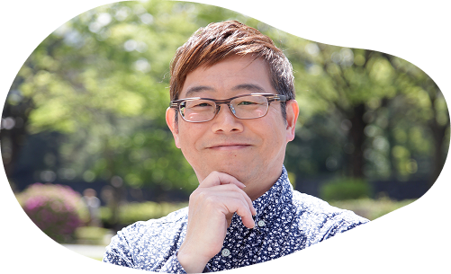 緑豊かな背景に、小崎先生が笑顔でこちらを見ている